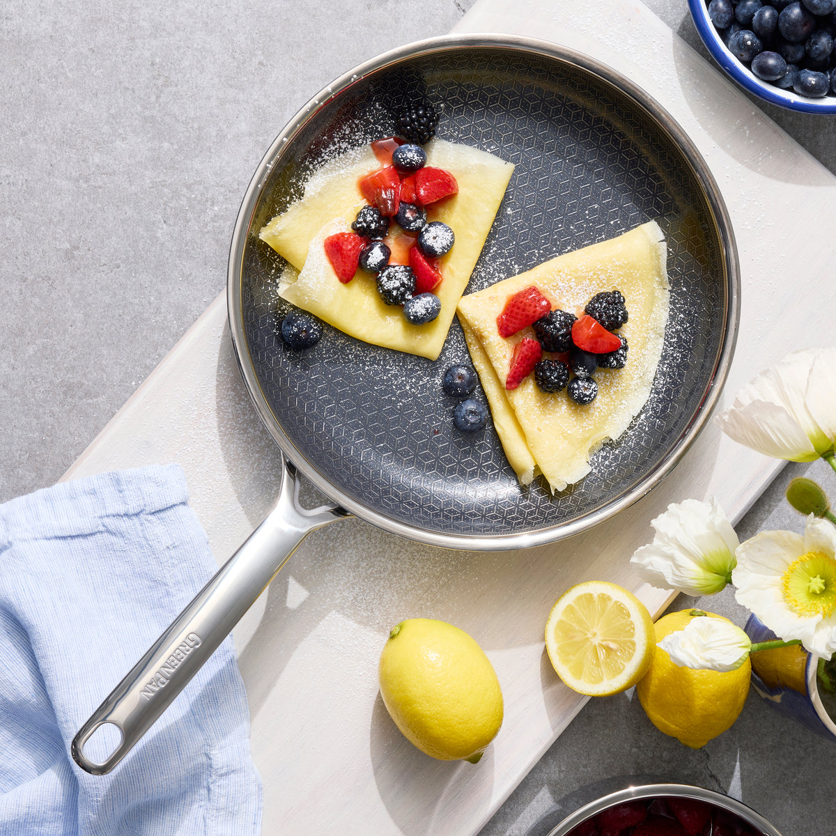 Two crepes with berries in a gray hybrid ceramic nonstick skillet, lemons and flowers on the side, blue napkin nearby.