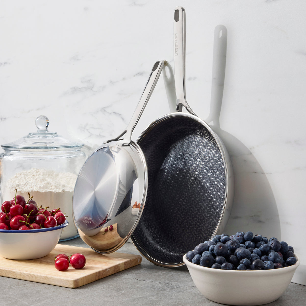 Two ceramic nonstick frypans, 10 and 12, hanging near bowls of blueberries, cherries, and flour.