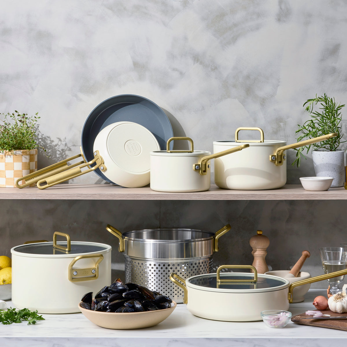 White 11-piece cookware set with gold handles, arranged with herbs, lemons, and kitchen items on a counter.
