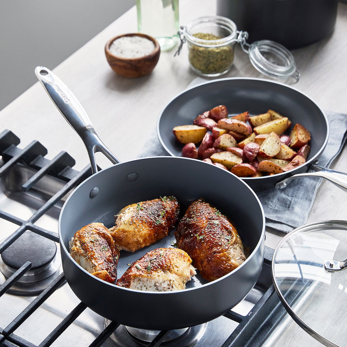 Two frying pans on a stove with food, surrounded by kitchen items.