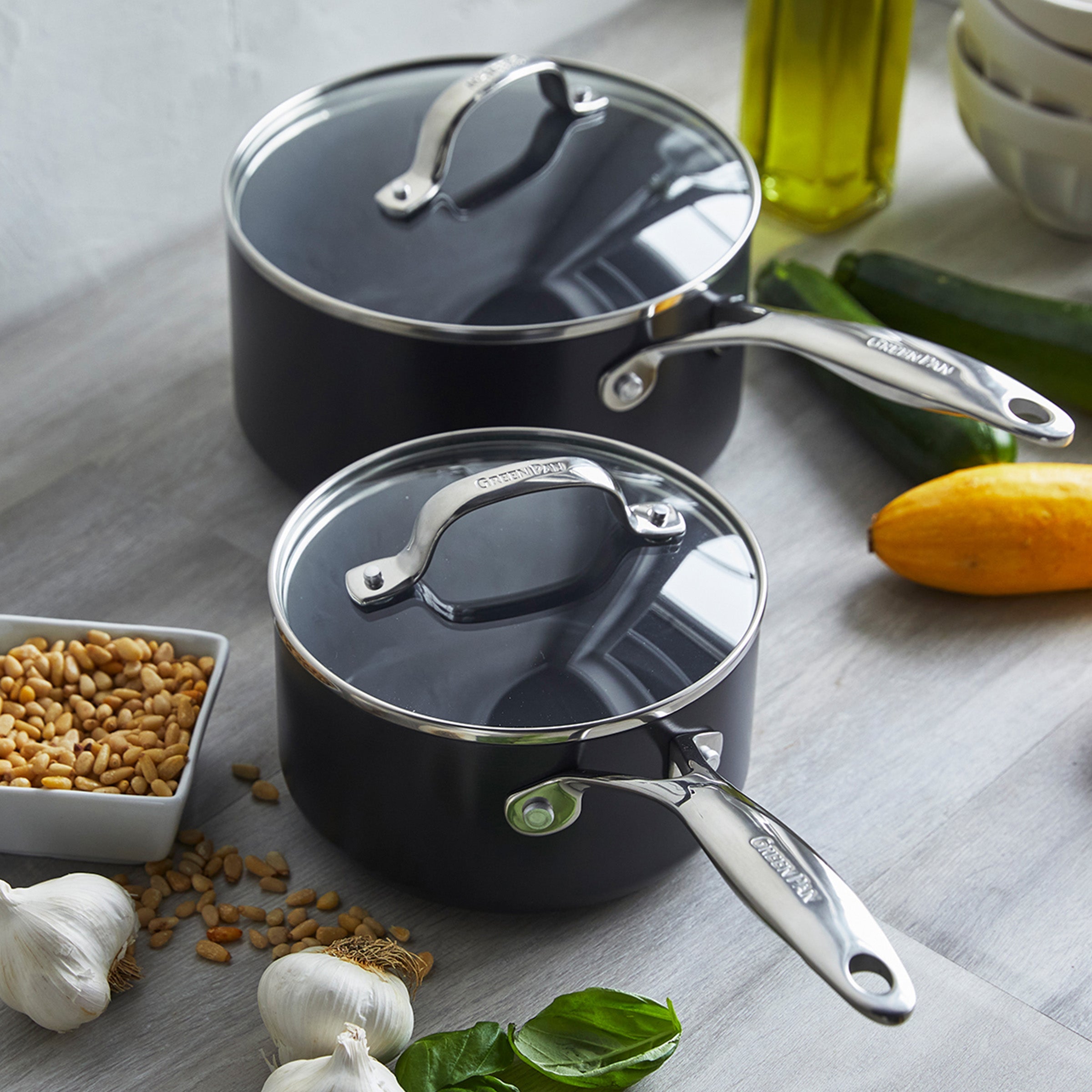 The Valencia Pro Ceramic Nonstick 1.5-Quart and 3-Quart Saucepan Set with Lids in gray sits on a kitchen counter among garlic, basil, pine nuts, olive oil, and squash, offering durability and scratch resistance for daily cooking.