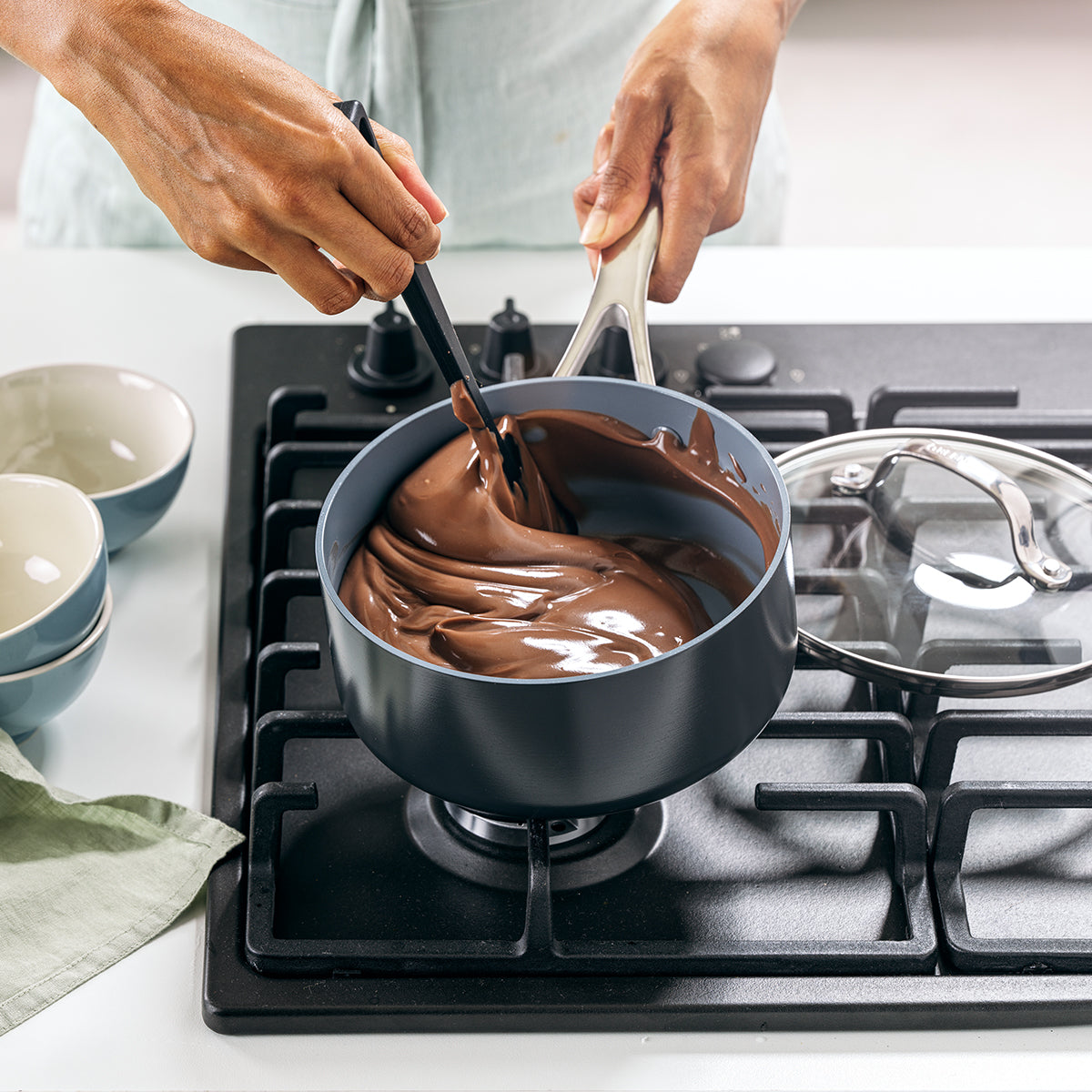 Someone stirs thick, melted chocolate in the Valencia Pro Ceramic Nonstick 1.5-Quart Saucepan on a stovetop, holding the scratch-resistant pan and spatula, with bowls and a green cloth nearby.