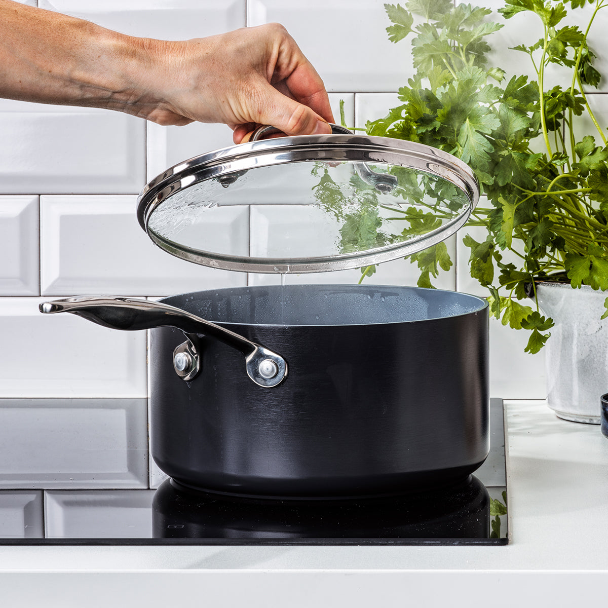 A hand lifts the glass lid of a Valencia Pro Ceramic Nonstick Saucepan (1.5-Qt or 3-Qt) as steam rises on an induction cooktop, with fresh parsley and white tile kitchen walls in the background.