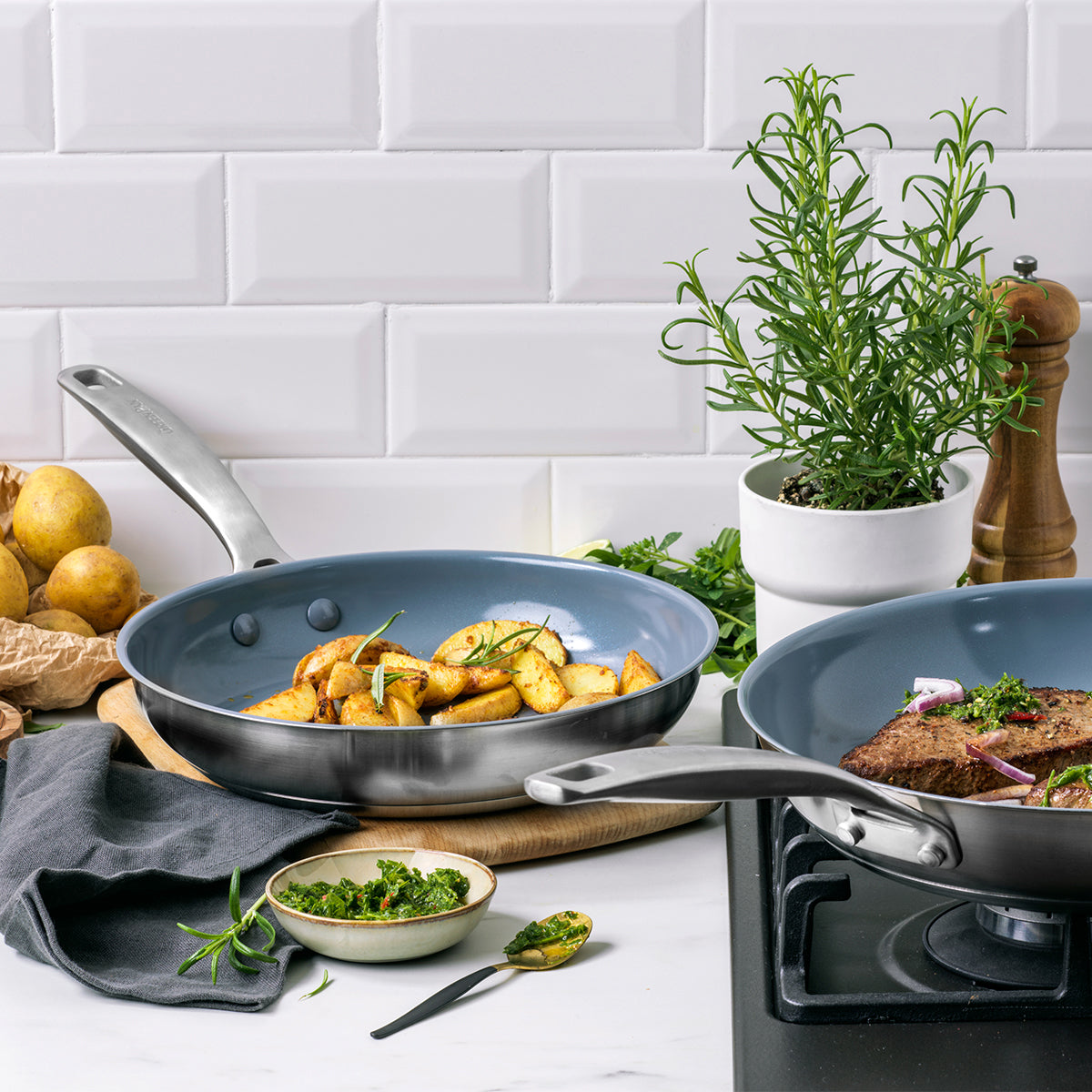 Stainless steel ceramic frypan set with roasted potatoes and seared meat on a white tile surface.
