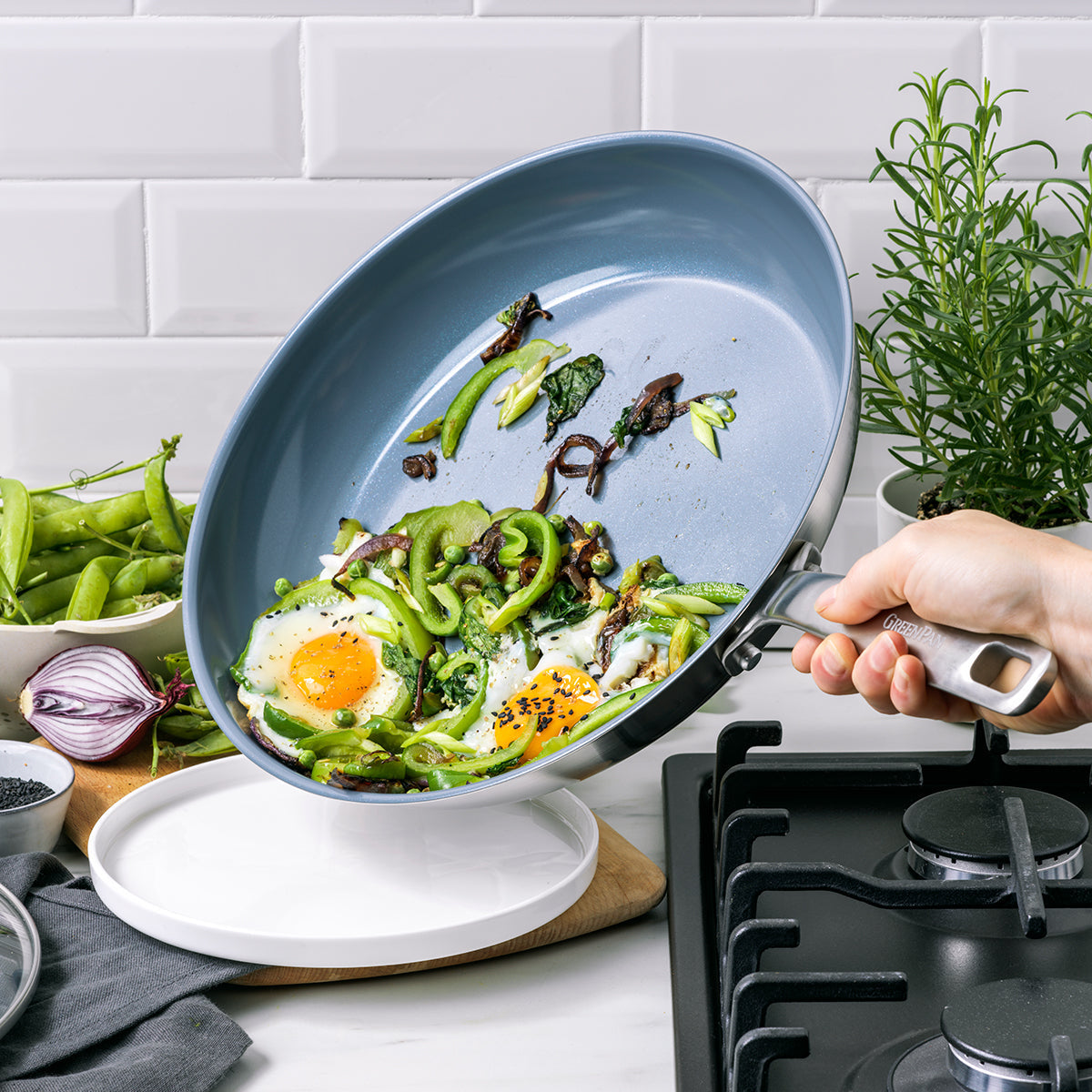 Hand holding a ceramic skillet with eggs and vegetables by the stovetop.