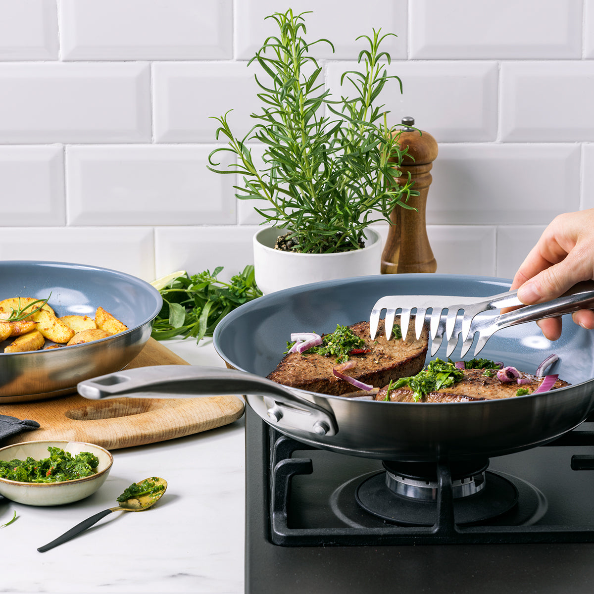 Steak is seared with tongs in a stainless steel nonstick pan, with herbs and potatoes visible in the background.