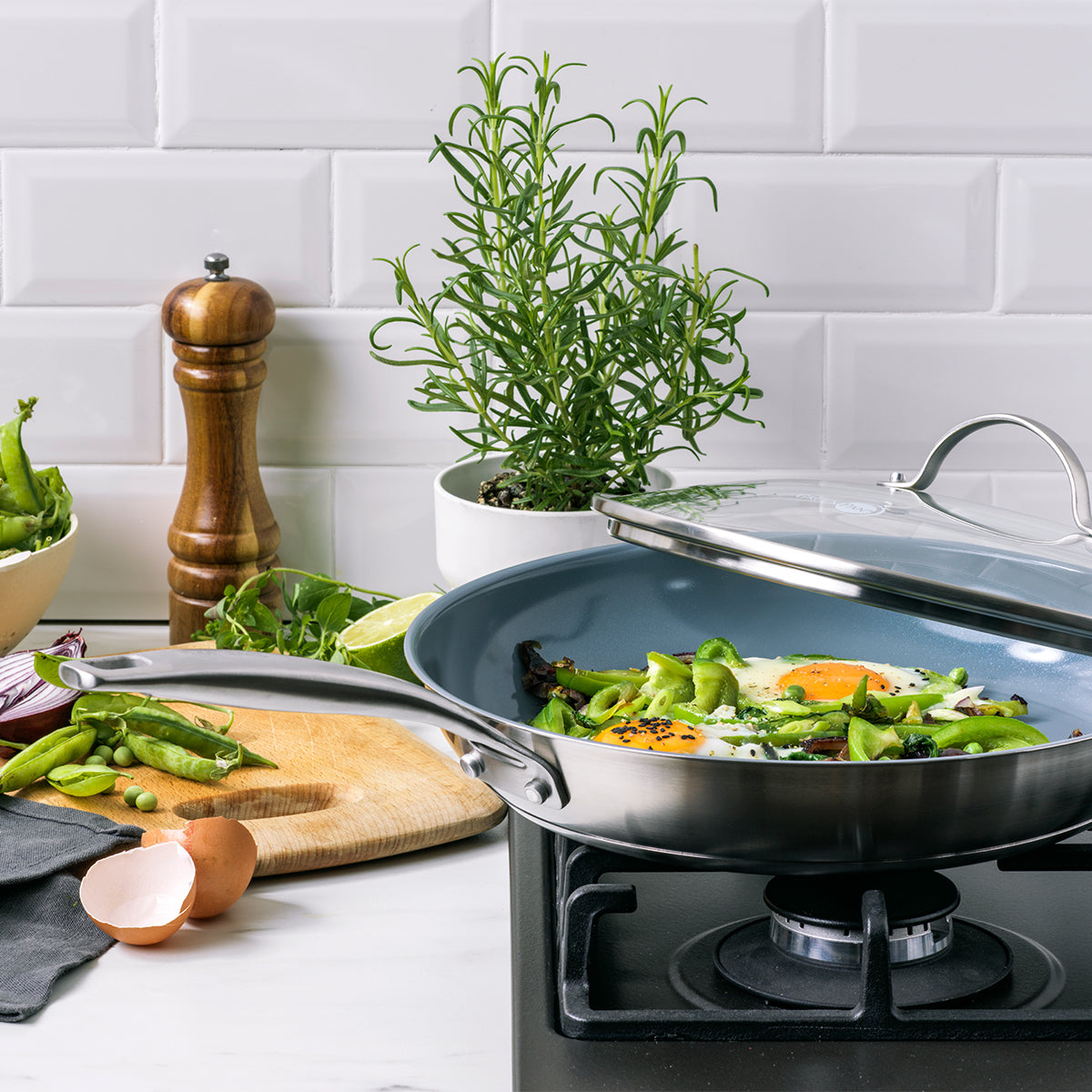 Ceramic nonstick stainless steel pan from 10-piece set on stove with eggs, vegetables, and herbs nearby.