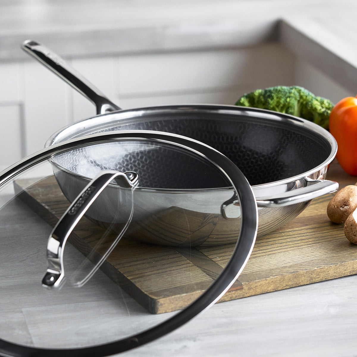 Hybrid ceramic nonstick 12 wok with lid on a cutting board, surrounded by assorted vegetables in a kitchen.