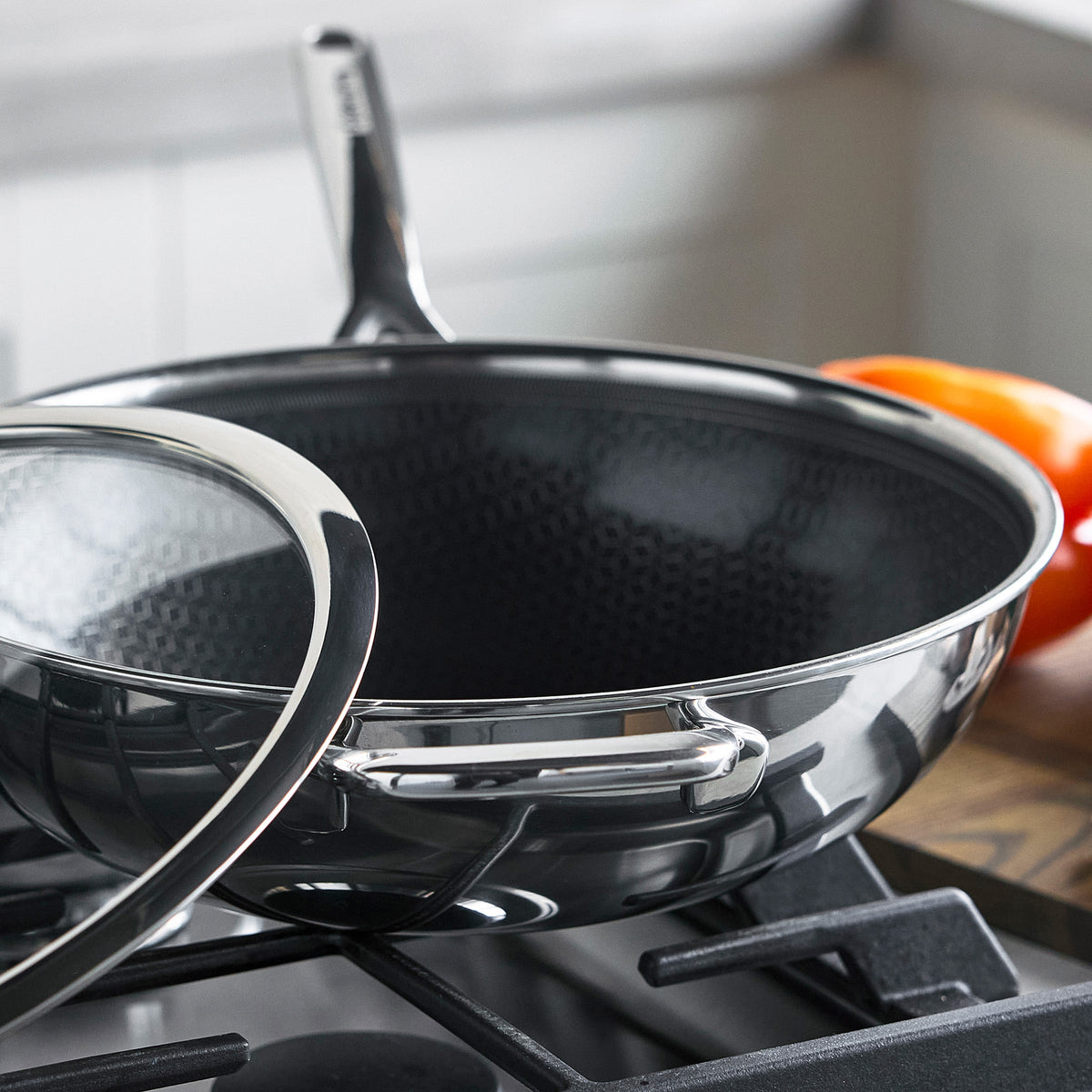 Black ceramic nonstick wok with lid on a gas stove, tomato visible in the background.