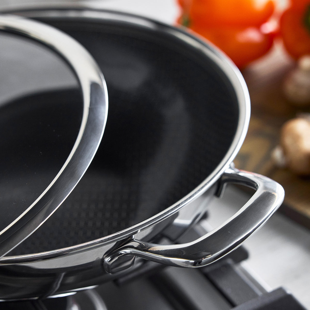 Hybrid ceramic nonstick wok with glass lid on a stovetop, red tomatoes in the background.