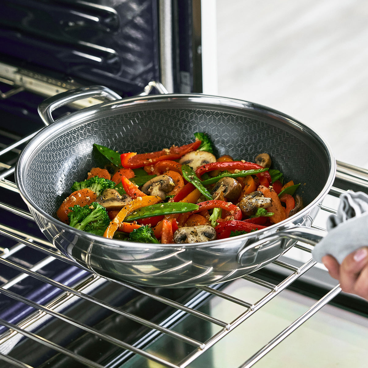 Gray hybrid ceramic nonstick wok with lid, filled with vegetables, on an oven rack being held by a mitt.