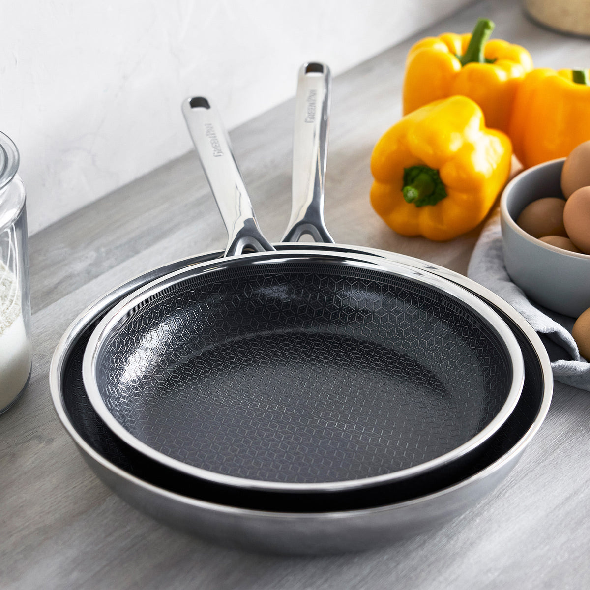 Two gray ceramic nonstick frypans, 10 and 12, stacked on a counter near peppers, eggs, and a glass of milk.
