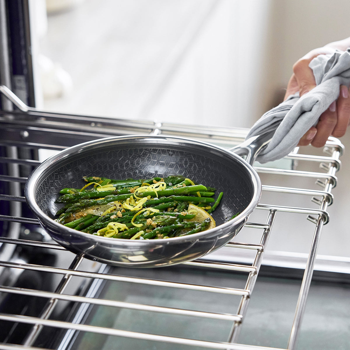 Hand holding a ceramic nonstick frypan with cooked asparagus and onions on an oven rack.