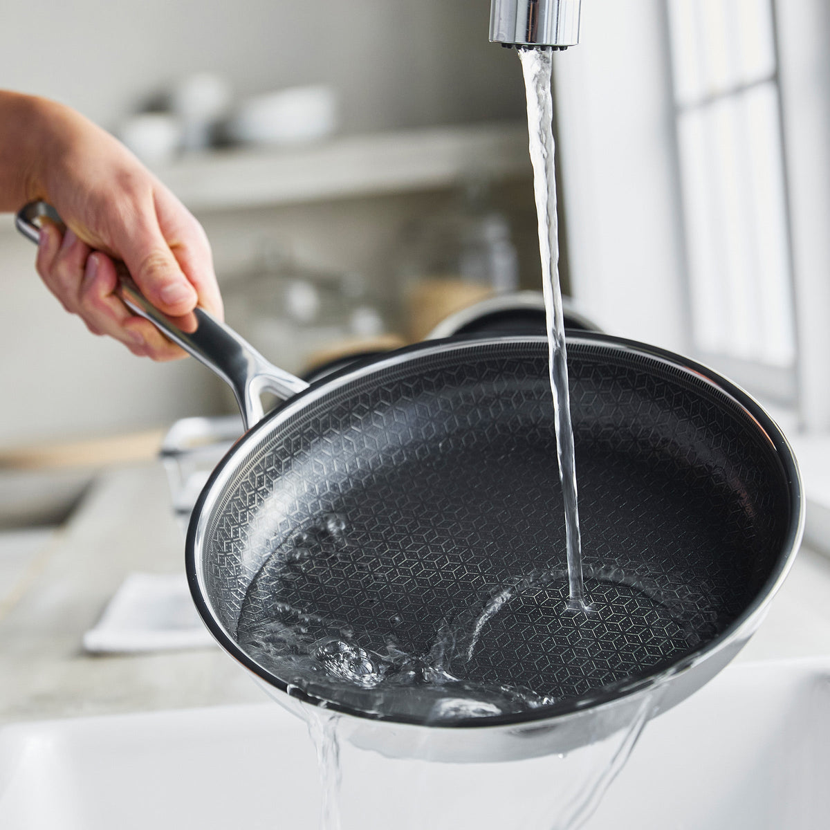 Hand rinsing a ceramic nonstick pan under running water in a kitchen sink.