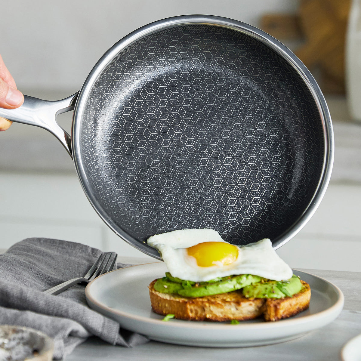 Hand holds a black ceramic nonstick pan above avocado toast with egg on a white plate.