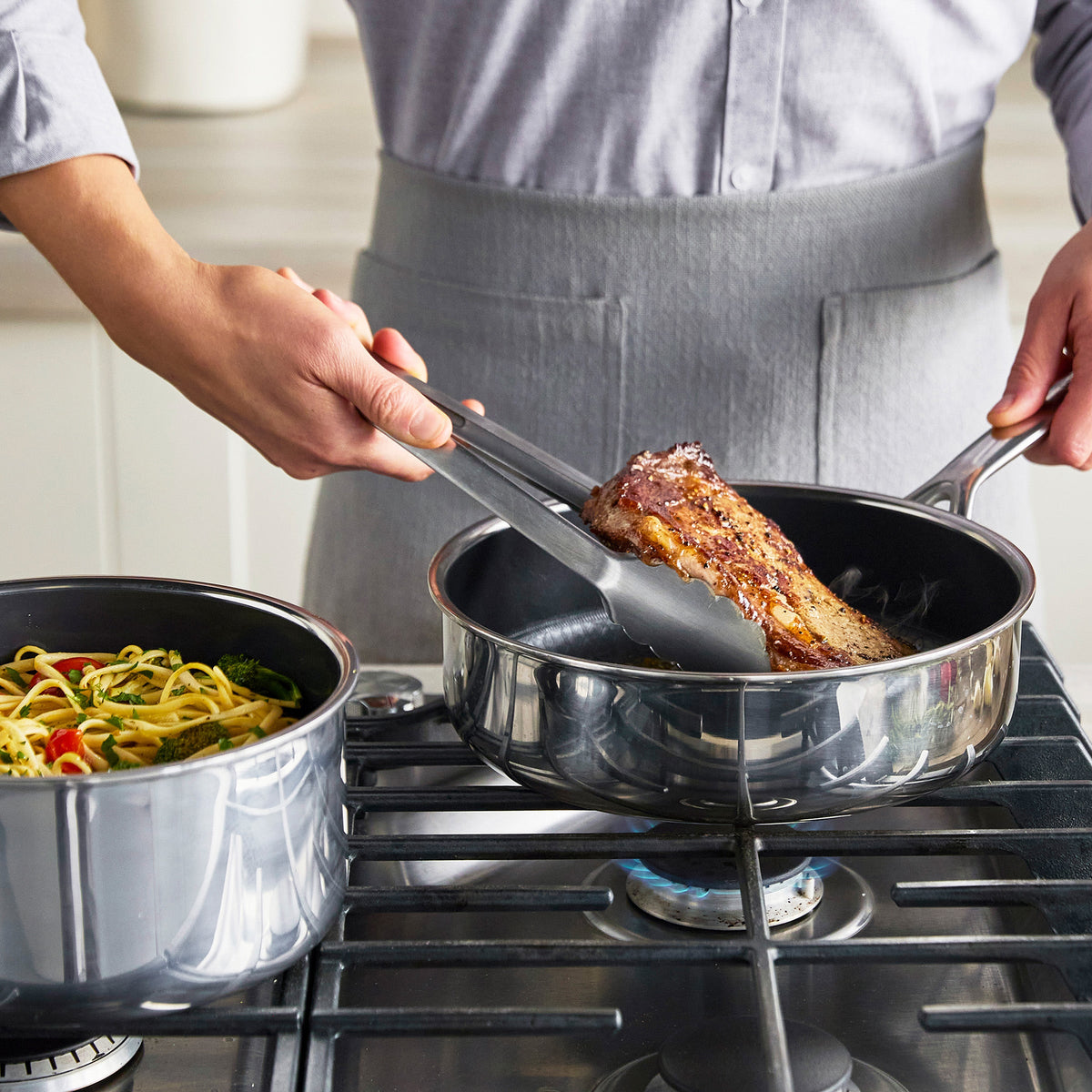 Steak cooking with tongs in a Spectra hybrid ceramic nonstick pan; vegetables and pasta visible in nearby pots.