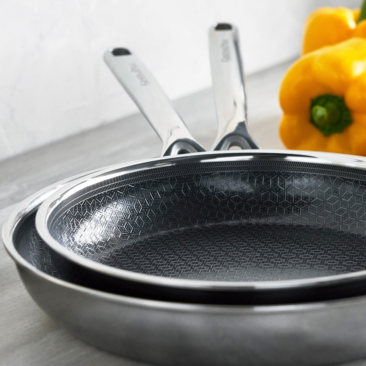 Stacked hybrid ceramic cookware set, 12 pieces, gray with metal handles, on counter beside yellow bell peppers.