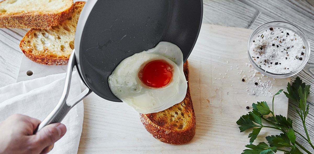 Fried egg sliding from skillet onto toast on white cutting board, with salt, pepper, and parsley beside.
