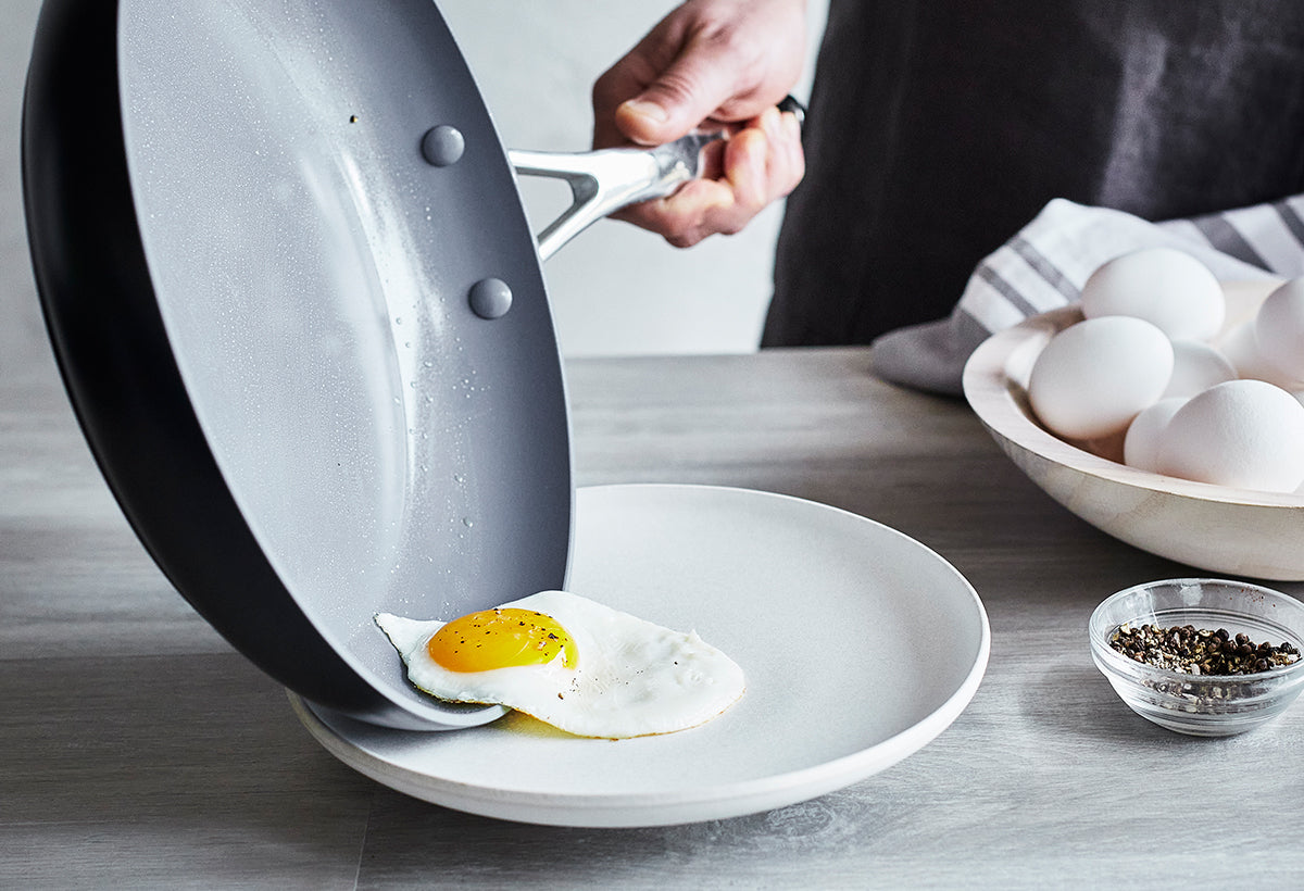 A fried egg is being slid from a GreenPan skillet onto a plate; whole eggs and a pepper grinder sit on the table.