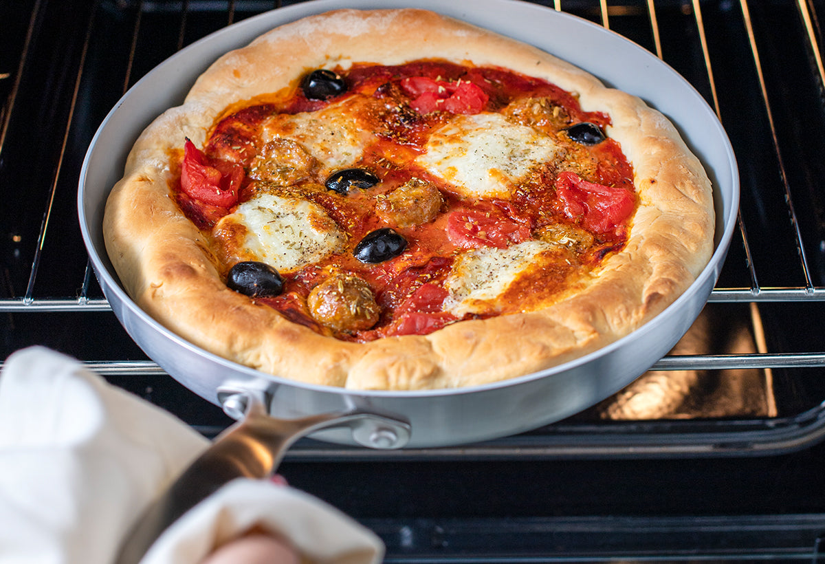 Homemade pizza with cheese, meatballs, tomatoes, and black olives in a pan being held in front of an oven.