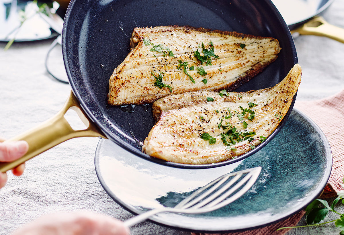 Cooked fish fillets with herbs in a GreenPan frying pan, served with a metal spatula onto a plate.
