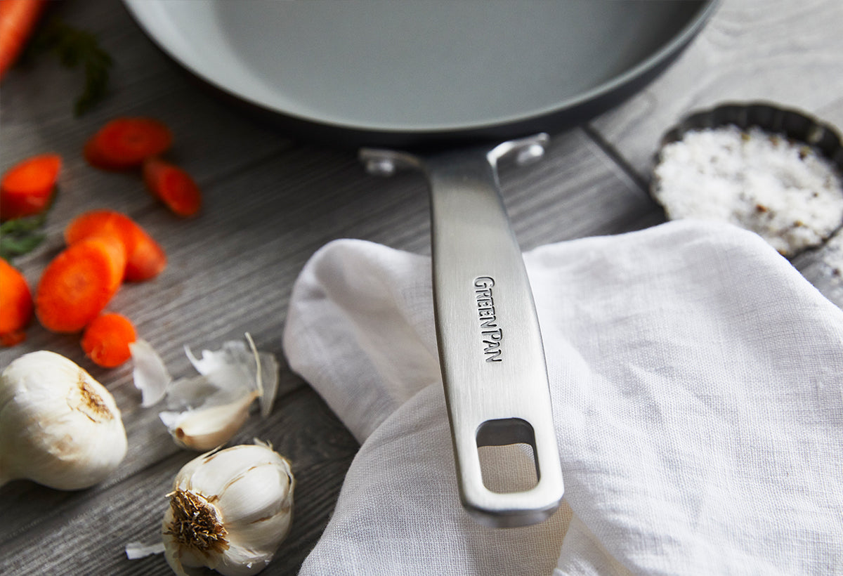 GreenPan skillet on a wooden table with garlic, carrot slices, salt, and a white cloth nearby.