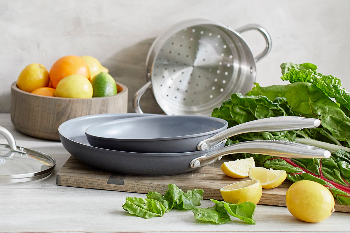 Two frying pans with lemons, leafy greens, a GreenPan skillet, and a metal strainer on a kitchen counter.