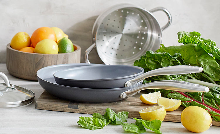 Set of two gray skillets with fresh lemons, leafy greens, citrus fruits, and a metal colander on a kitchen counter.