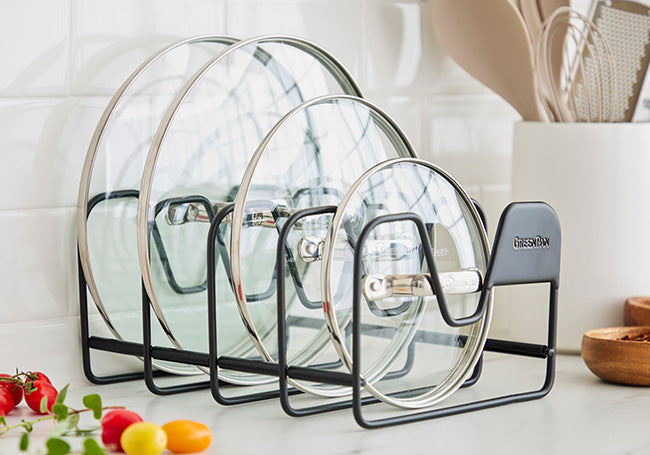 Black metal rack holding four glass pot lids upright on a kitchen counter with utensils and vegetables nearby.