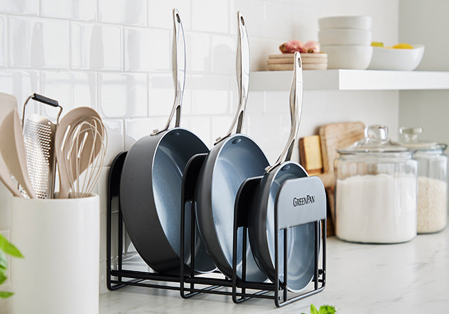 Set of three ceramic nonstick skillets in a rack on a kitchen counter with surrounding utensils, jars, and dishes.