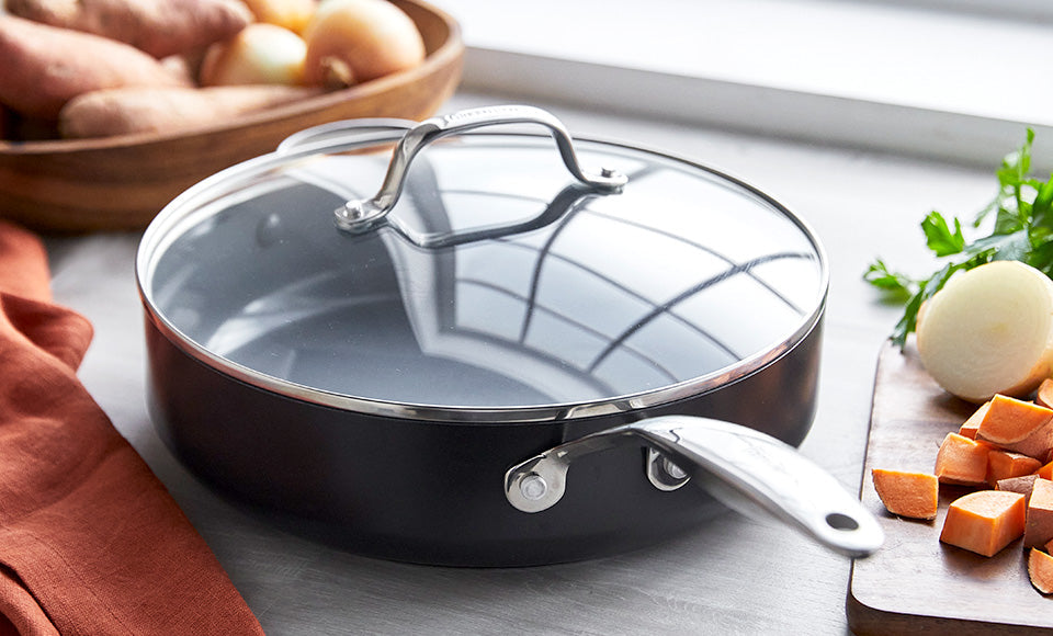 Ceramic nonstick skillet with metal handle on countertop, next to chopped onions, sweet potatoes, and other vegetables.