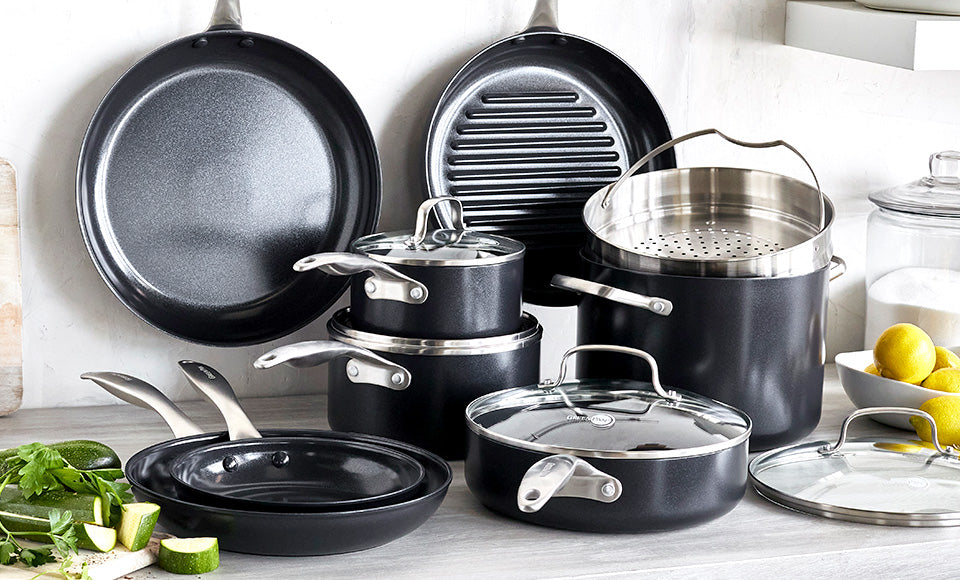 Black GreenPan cookware set with pots, pans, and lids on a kitchen counter with assorted vegetables and lemons nearby.