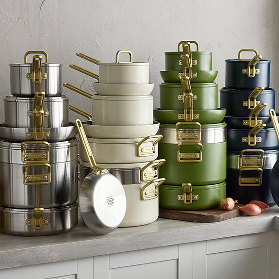 Ceramic cookware set in silver, white, green, and navy with gold handles displayed on a kitchen counter.