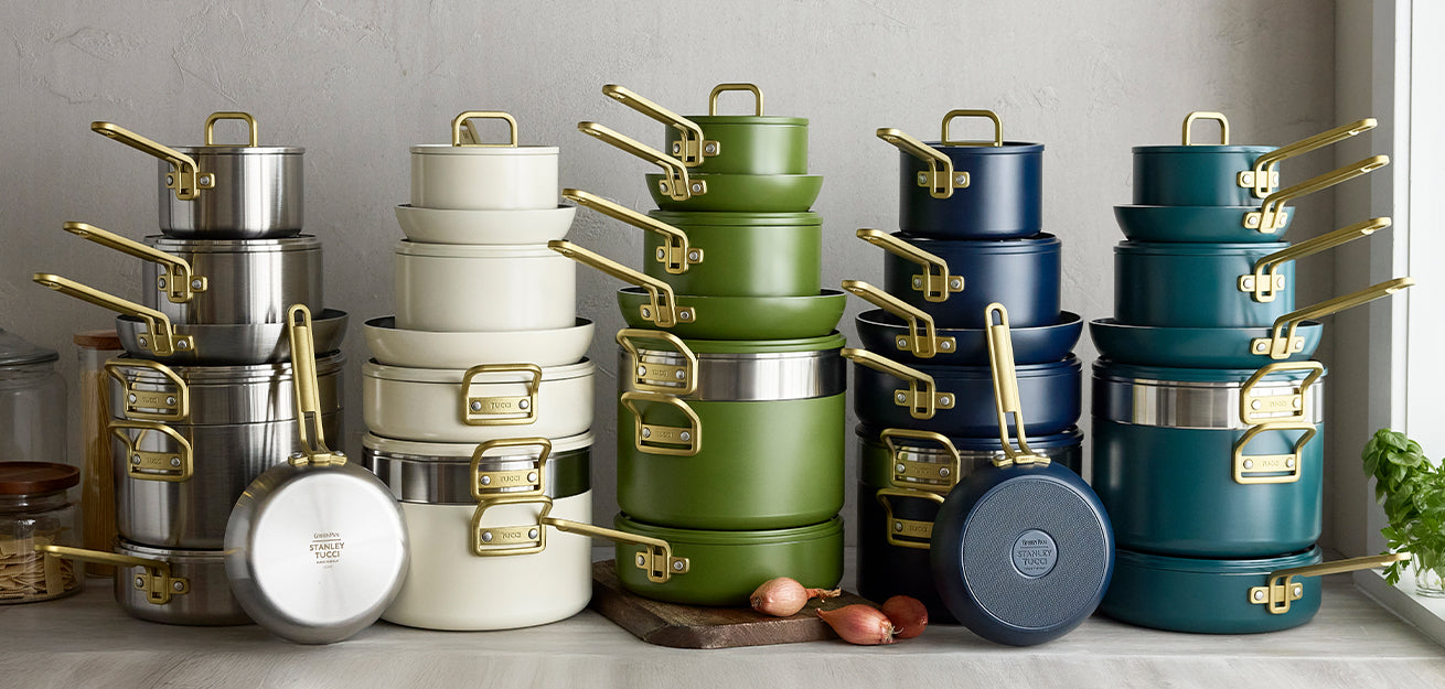 Stacked cookware set in green, cream, navy, teal, and stainless steel with gold handles on a kitchen counter.