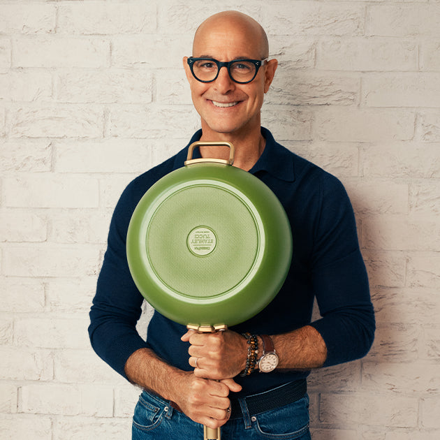 Man in glasses and navy shirt holding a single green ceramic nonstick skillet vertically against a white brick wall.