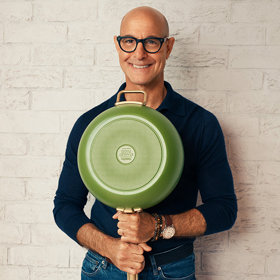 Man with glasses holding a large green skillet upright against his chest in front of a white brick wall.