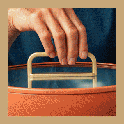 Steam rises as a hand lifts the lid from an orange GreenPan pot.