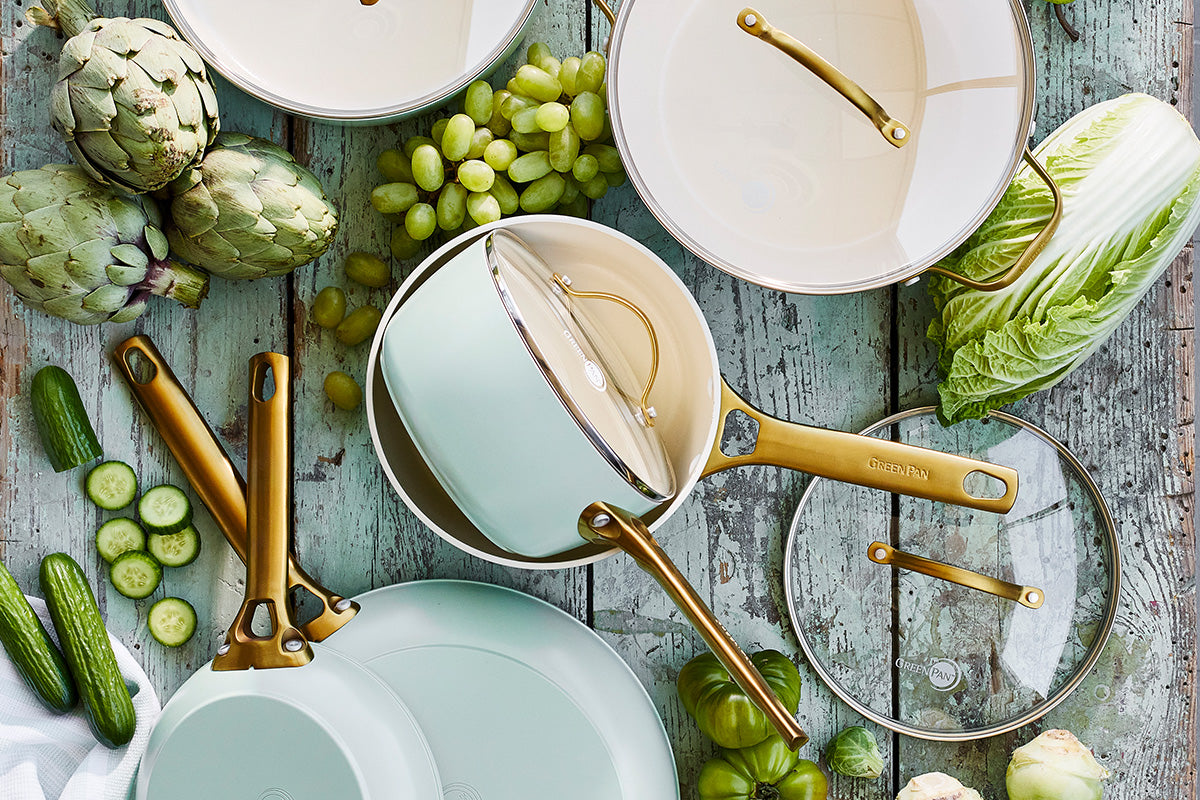 Cookware set with gold handles displayed on a rustic blue table, surrounded by artichokes, grapes, lettuce, cucumbers, and tomatillos.