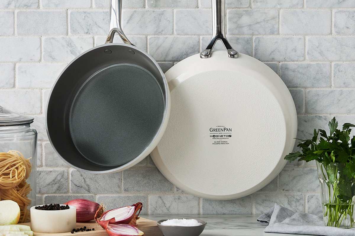 Set of two ceramic nonstick pans, one hanging to show the interior, on a kitchen counter with pasta, herbs, shallots, and salt.