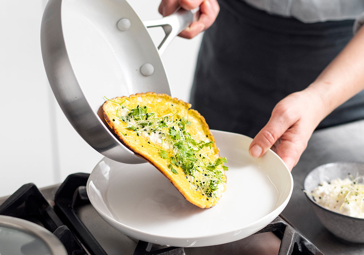 Cooked omelet with greens being slid from a black skillet onto a white plate on a kitchen counter.