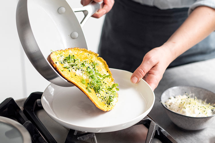 GreenPan ceramic nonstick pan with a cooked herb omelette being slid onto a white plate, fresh toppings nearby.