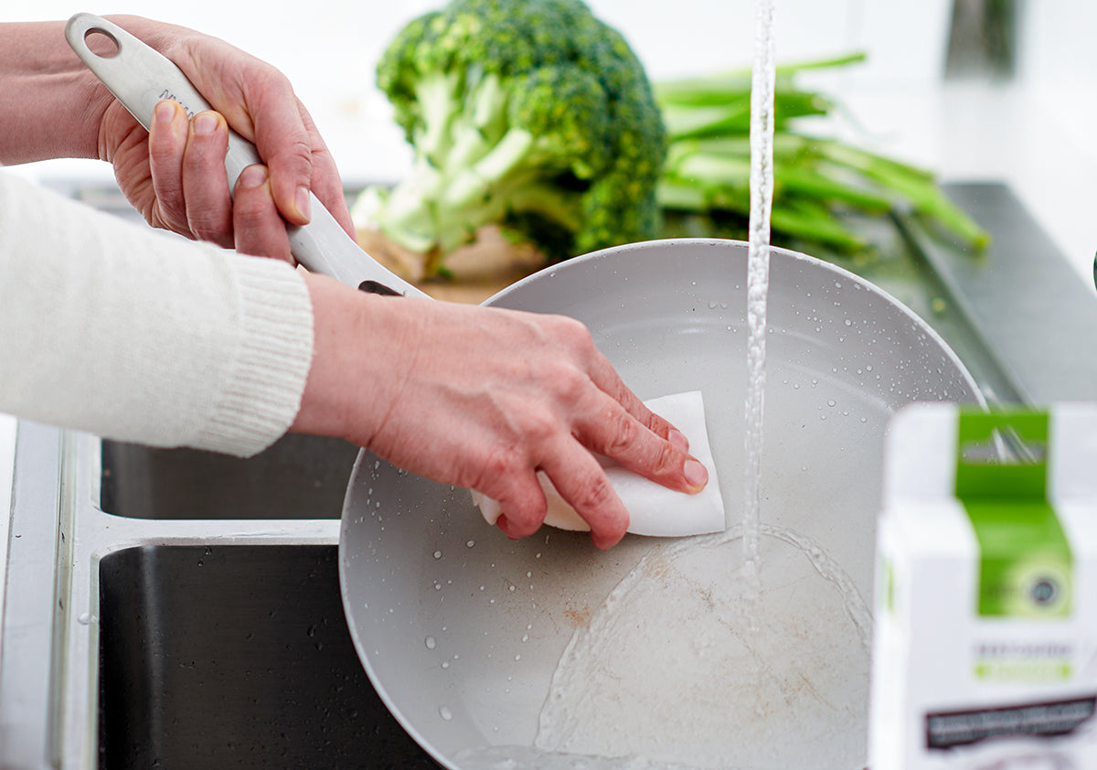 Hand cleaning a white ceramic nonstick skillet in a kitchen, with broccoli and green onions on the counter nearby.