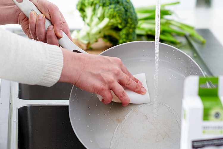 Hand washing a ceramic nonstick skillet under running water, fresh broccoli on a cutting board in the background.