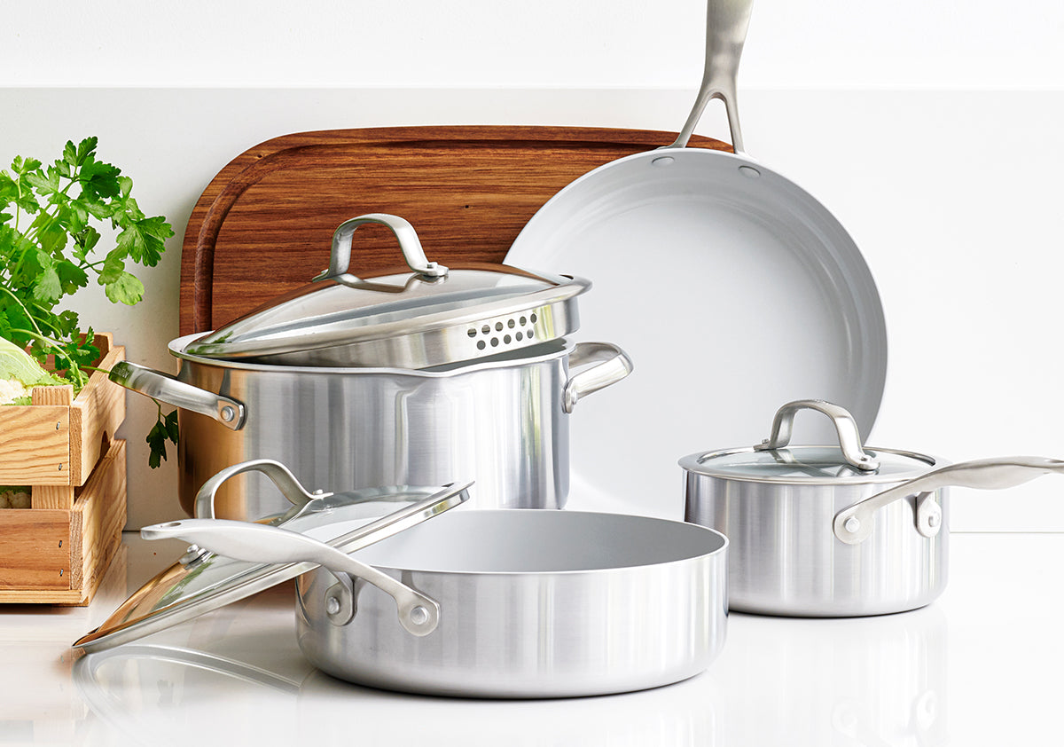 Green ceramic nonstick pots and pans with lids on a white counter, wooden cutting board and fresh herbs behind.