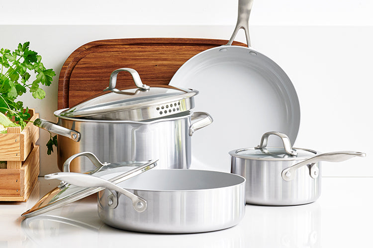 Silver cookware set with lids, includes pans and a skillet, on a white counter beside a wooden cutting board and fresh parsley.