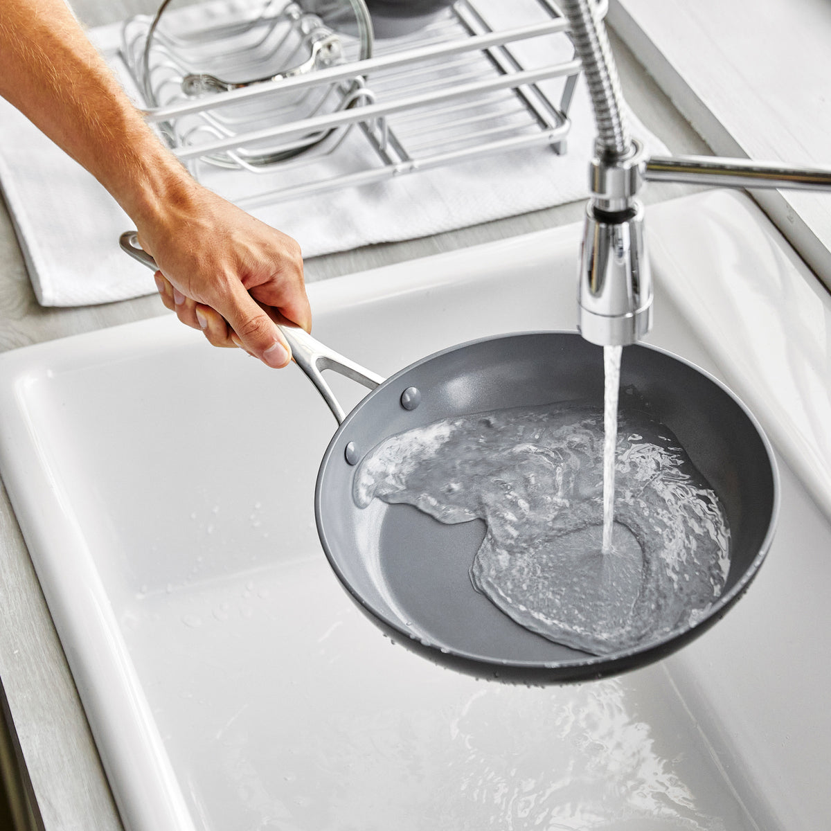 Hand rinsing a ceramic nonstick frying pan from a 13-piece set under running water in a kitchen sink.
