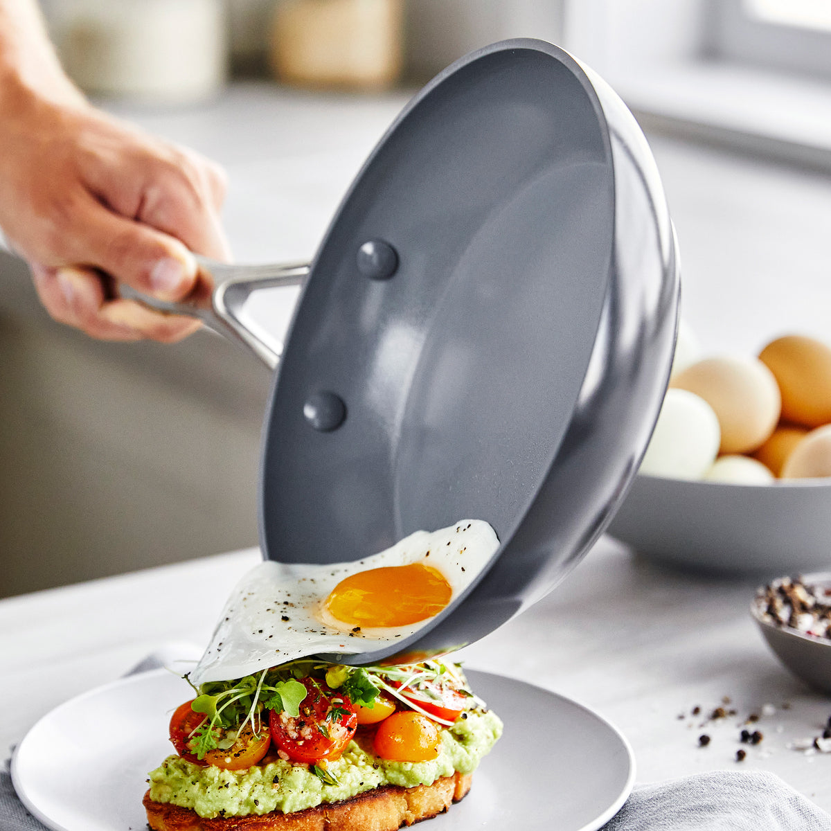 A hand sliding a sunny-side-up egg from a gray ceramic nonstick frypan onto avocado toast.