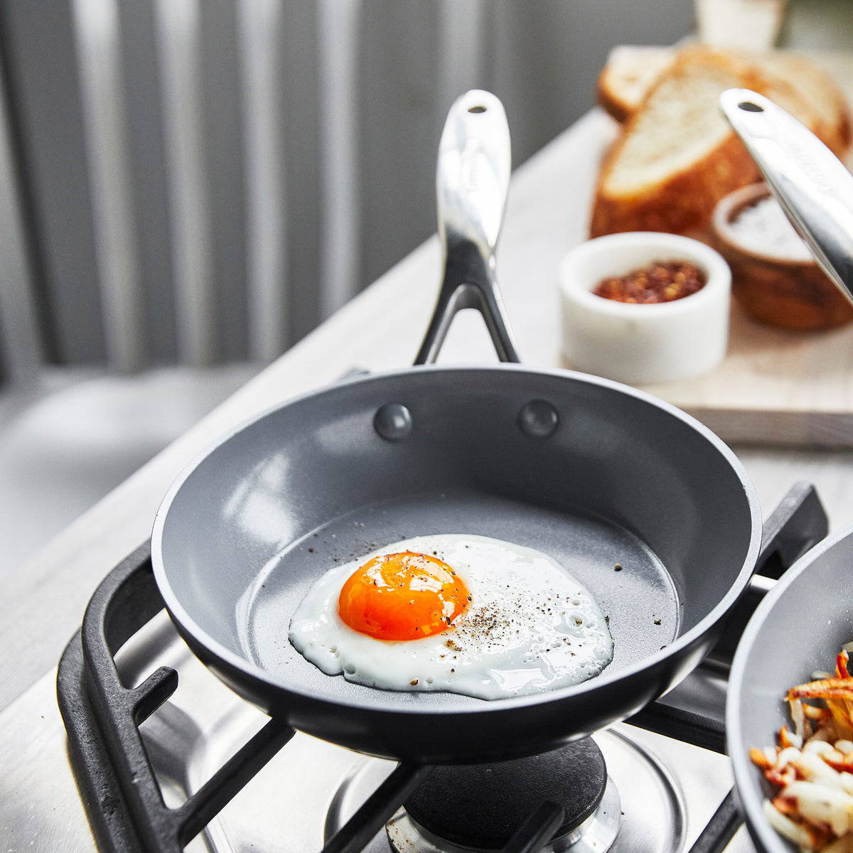 Sunny-side-up egg cooking in an 8-inch ceramic nonstick frypan with bread and spices blurred in the background.