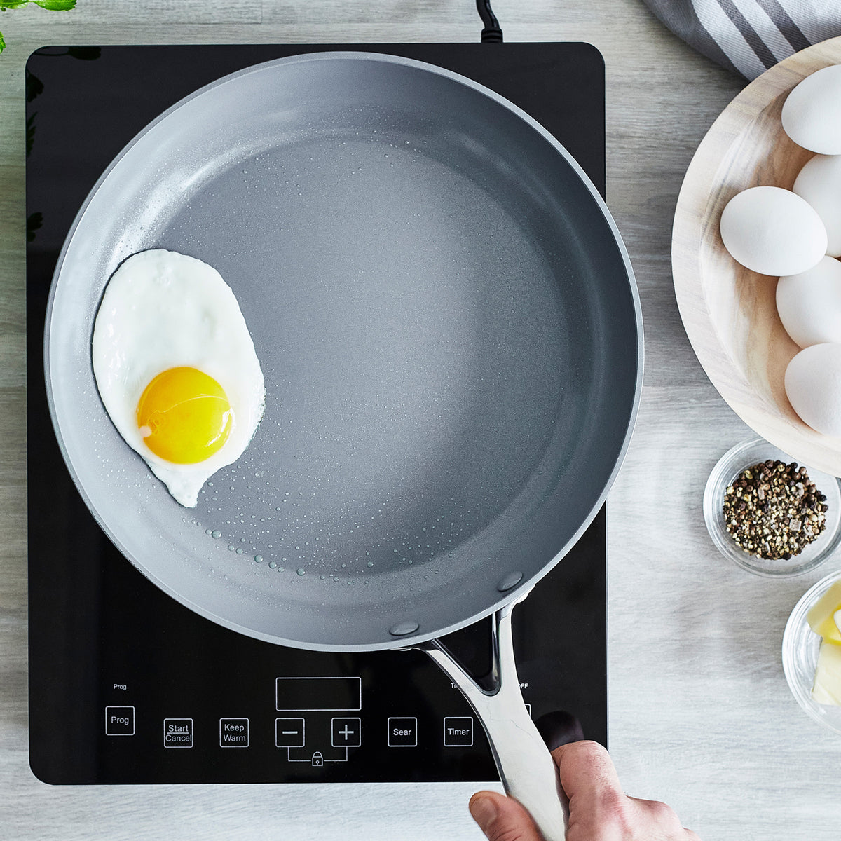 Hand holding a ceramic nonstick frypan with a sunny-side-up egg on an induction cooktop.