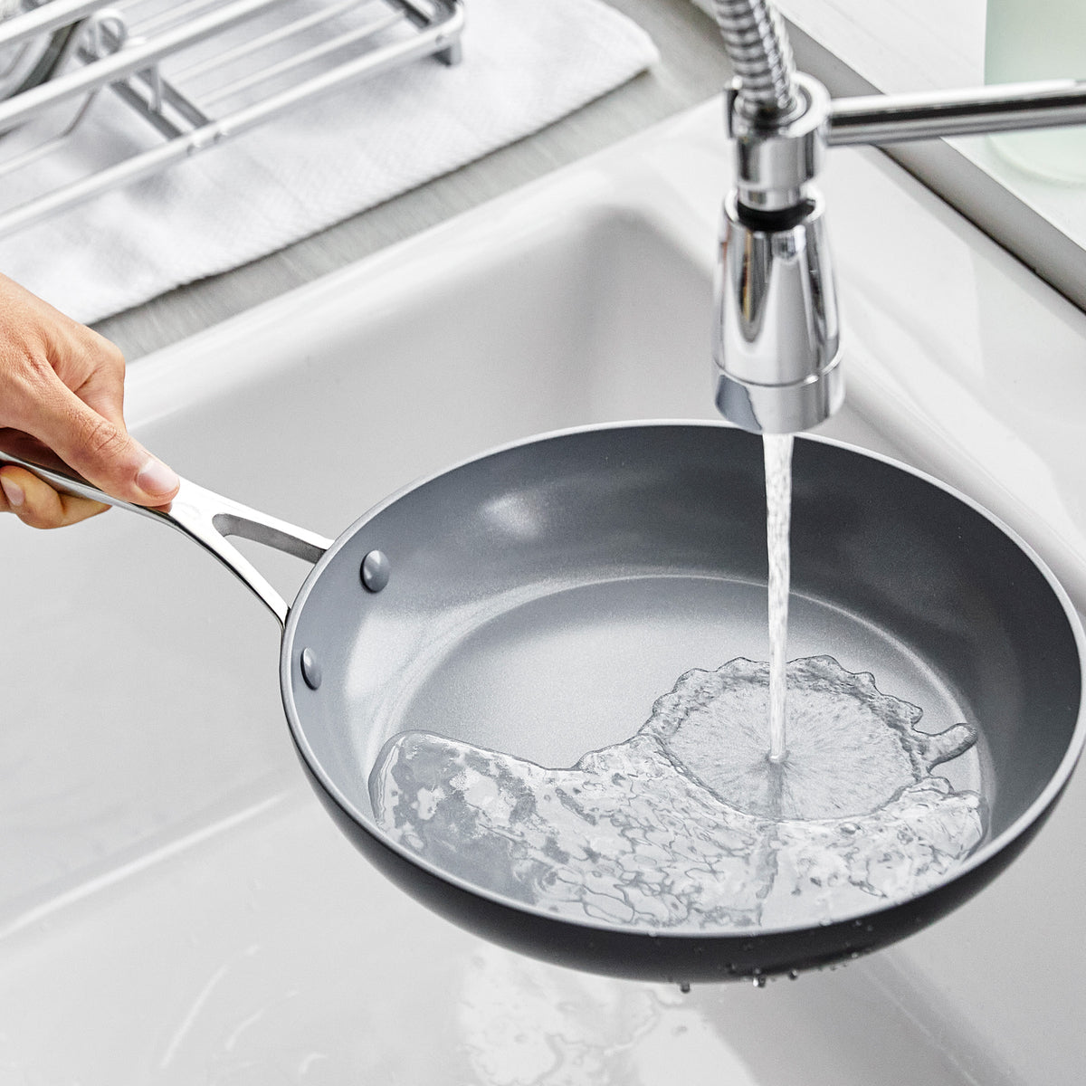A hand rinsing an 8-inch ceramic nonstick frypan under running water in a kitchen sink.